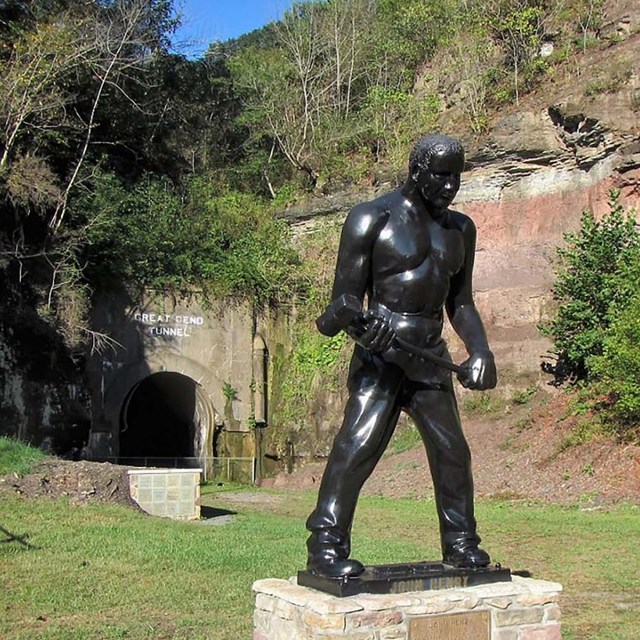 A black statue of a man holding a sledgehammer sits to the side in front of a railroad tunnel