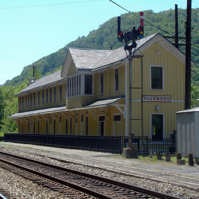 A long yellow wooden building with a sign that says Thurmond on it sits next to railroad tracks
