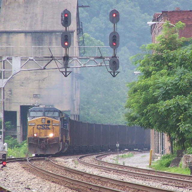 A blue and yellow CSX locomotive hauling coal cars on tracks through an old town past old buildings