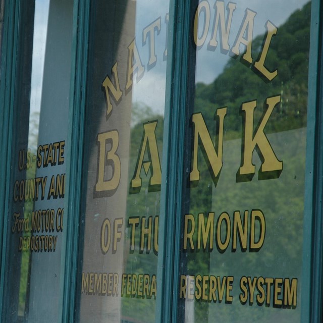 A window with a green wooden frame. The window has words on it that says National Bank of Thurmond.