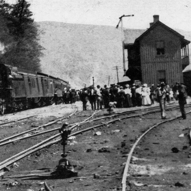 A picture from the early 1900s of a steam locomotive in front of a depot with people around it