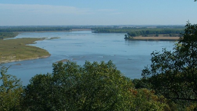 Wide flat river winding through a grassy landscape, with trees in the foreground