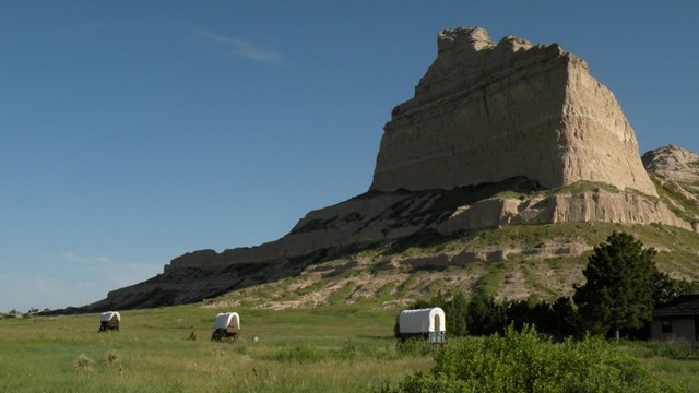 tree covered wagons in front of a very large rocky bluff in an otherwise grassland landscape