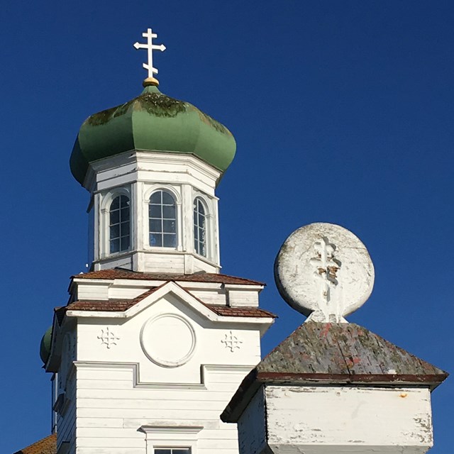 A photo of the Orthodox cross on a fence post with the church's green roofed dome in the background.
