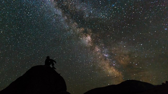 A man in silhouette against a starry night sits on a rock and contemplates the Milky Way.