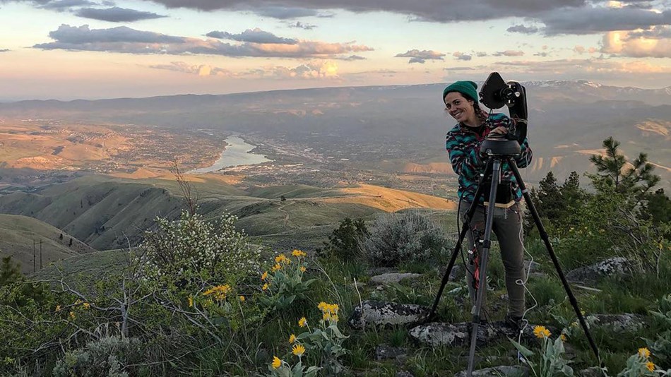 Sweeping valley view from atop a mountain shows a female scientist beside a tripod and CCD camera