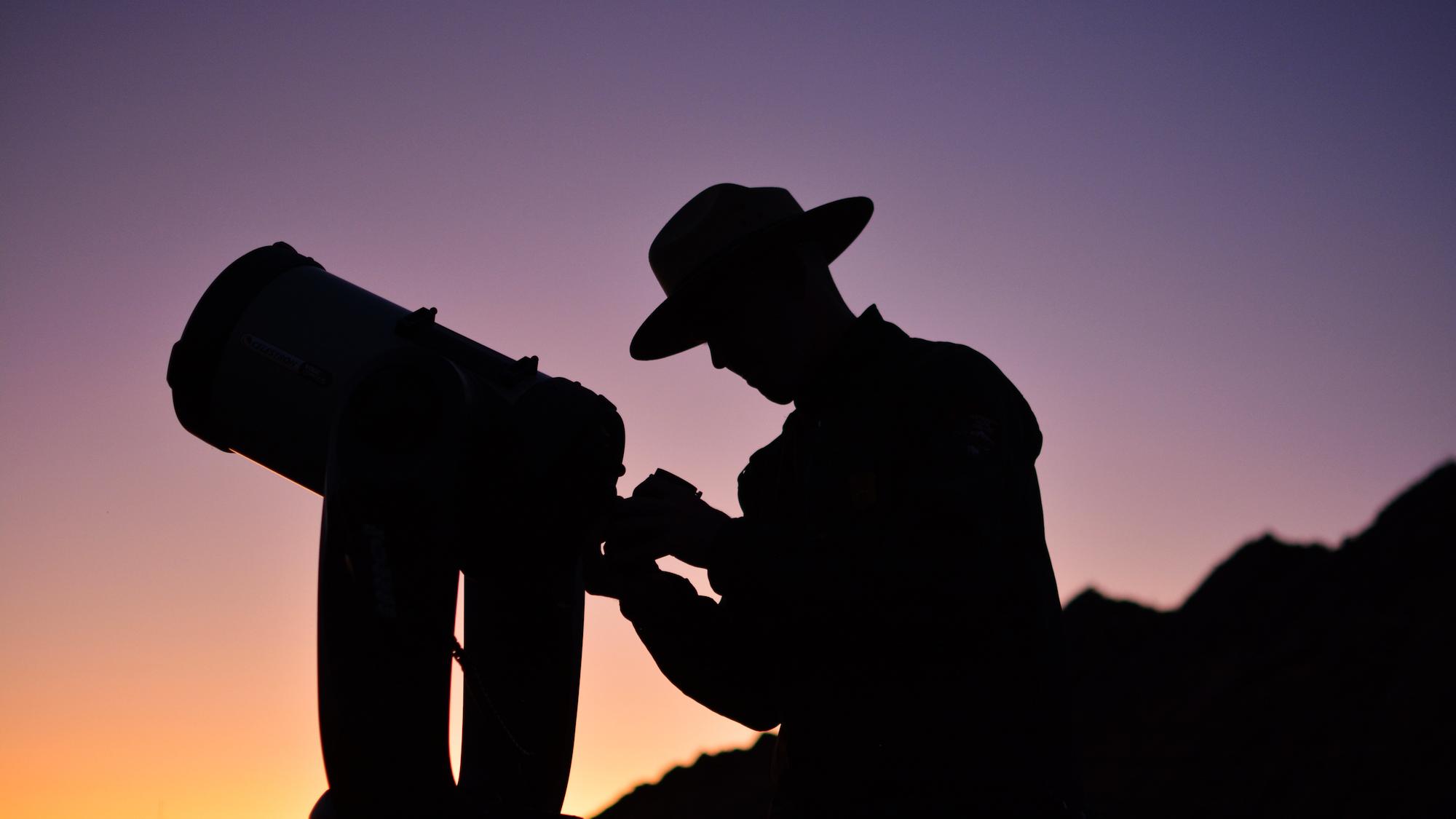 a silhouette of a ranger setting up a telescope at dusk.