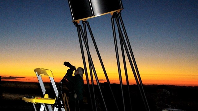 A man viewing the sky with binoculars stands next to telescope equipment