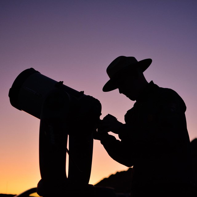 a silhouette of a ranger setting up a telescope at dusk.