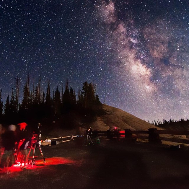 a group of people with telescopes and red lighting look up at a star filled sky and the milky way