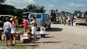 People and cars in a gravel parking lot with coolers and canoes.