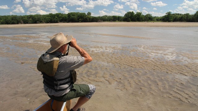 A man sits on the point of a canoe on a sandbar looking through binoculars.