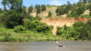 A child paddles a kayak upstream with a reddish bluff towering behind him.