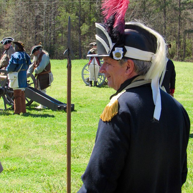 A man in a dark blue coat and a black hat with a red feather watching cannons