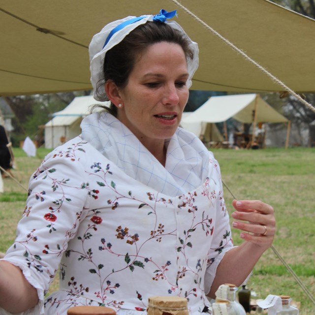 A woman in a white flowered gown and white cap at a table.
