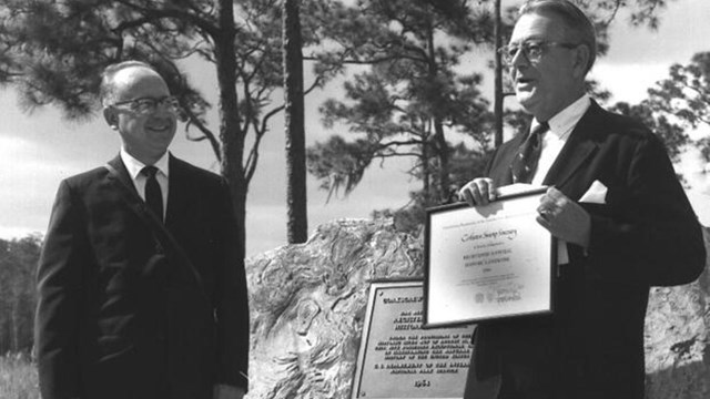 Two men pose next to a plaque with trees in background