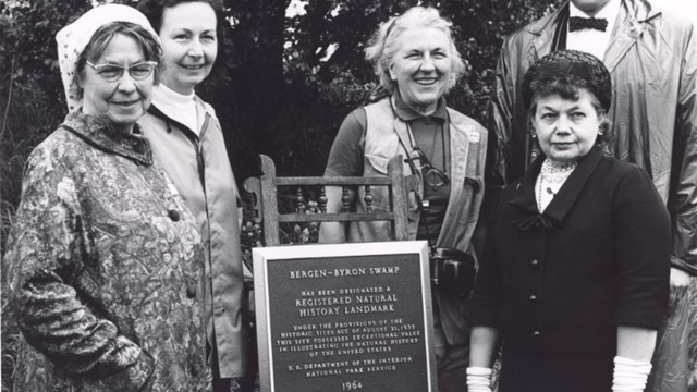 black and white photo of five people posed around a bronze plaque