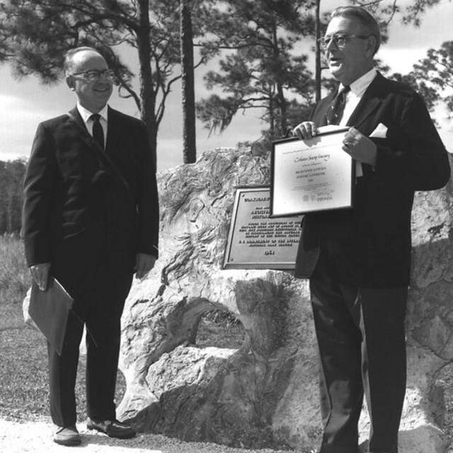 Two men pose next to a plaque with trees in background