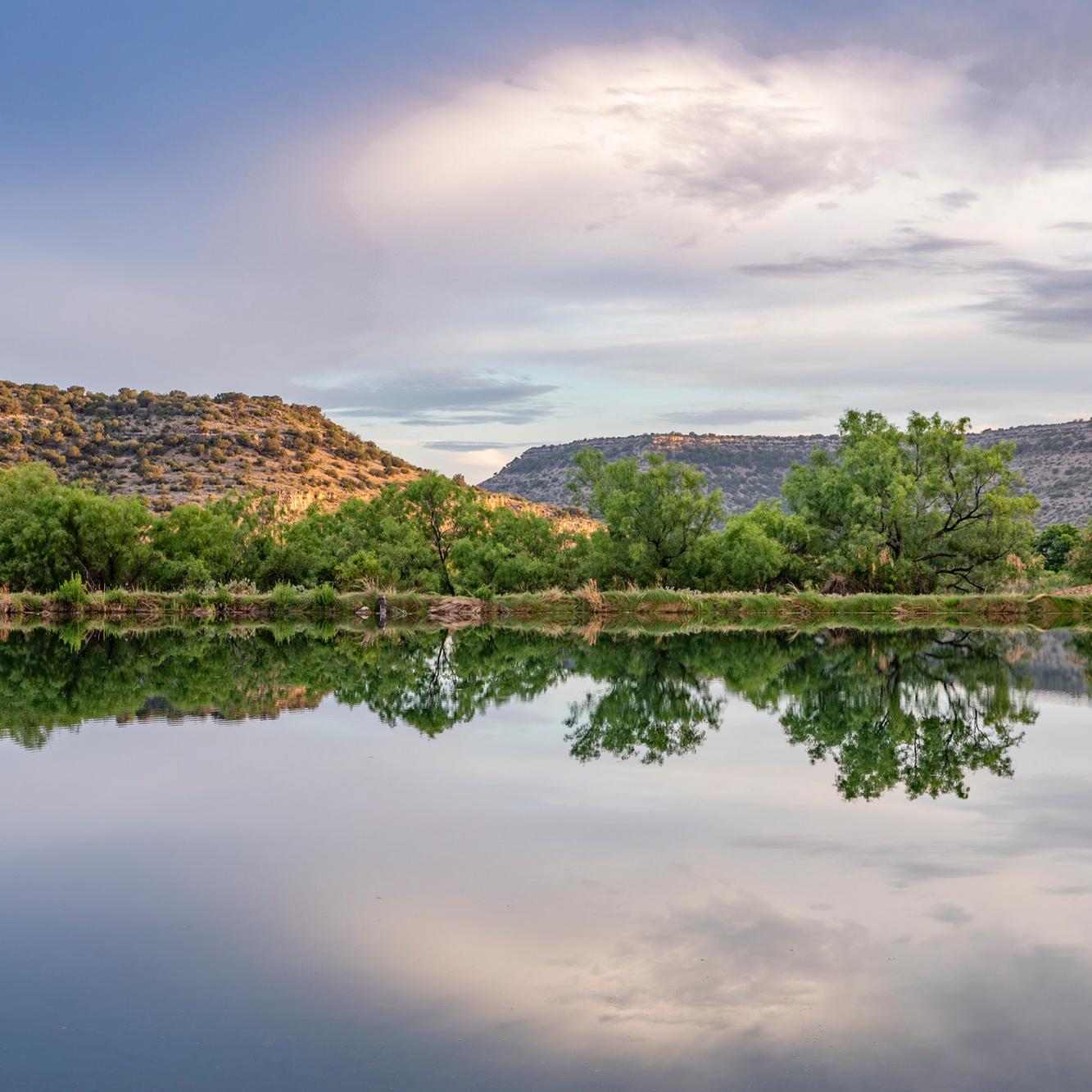 Smooth lake surface in foreground with trees, mountains, and sky in background
