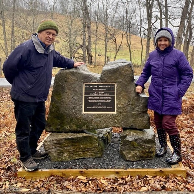 a man and a women stand next to an NNL designation plaque in a fall forest