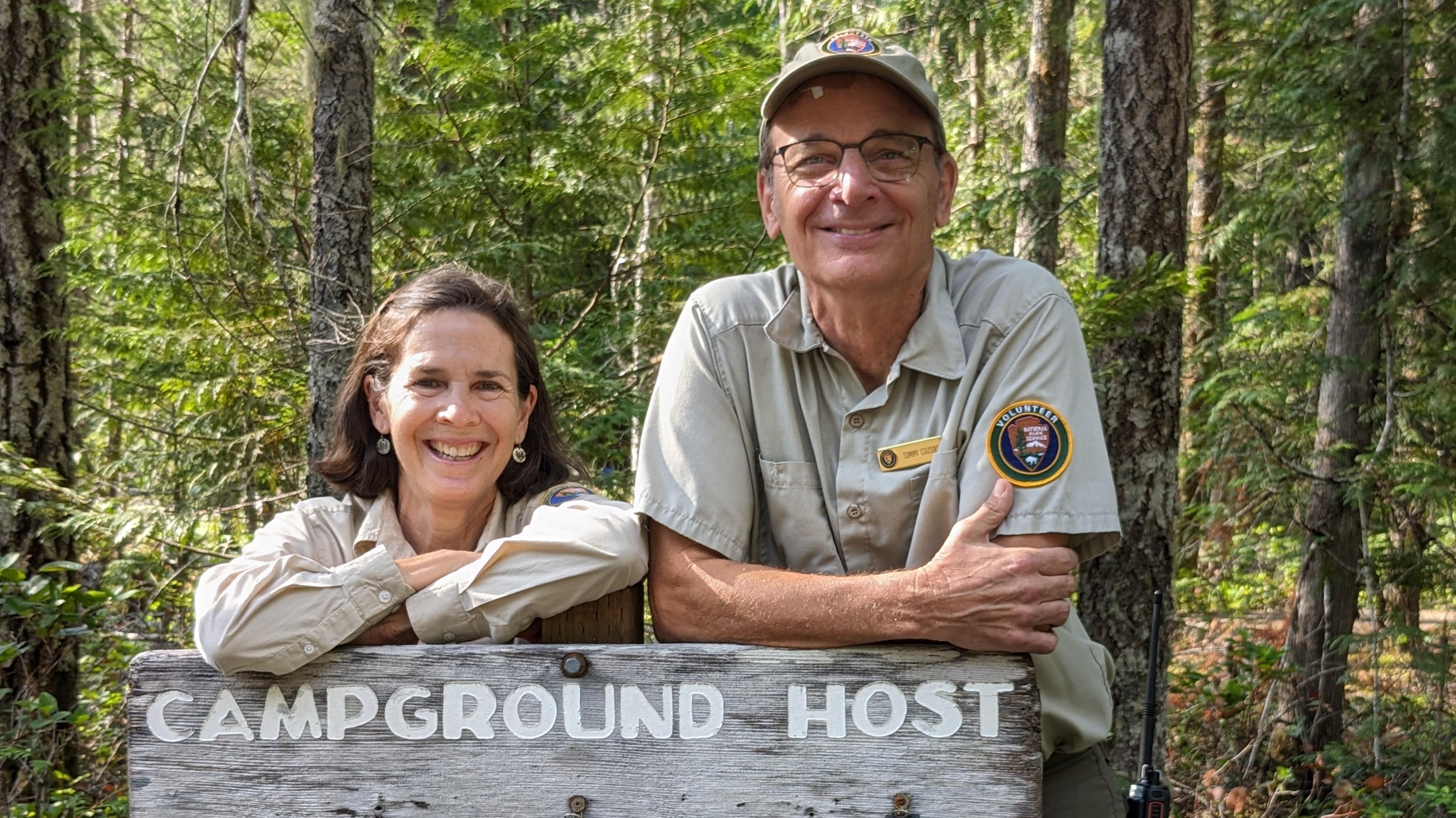 Two smiling volunteers with a sign that reads Campground Host with forest behind them.