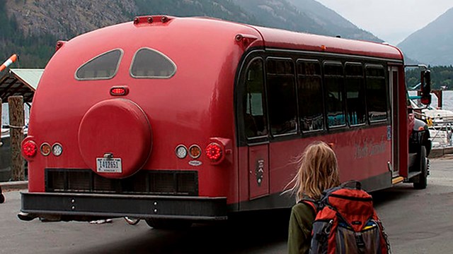 A hiker with a backpack stands near a long, red bus. 