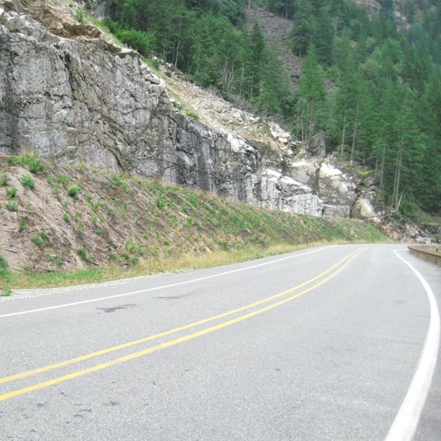 A two-lane road curves along a steep, rocky embankment.