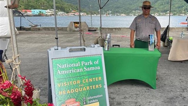 Outdoors; person in uniform standing behind table under tent with A-frame and flowers in front.