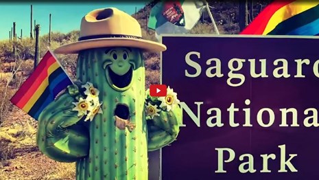 Cactus-shaped park ranger mascot holding a rainbow flag