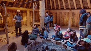 Ranger talking to a group of kids seated inside an earth lodge