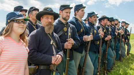 Child wearing a military hat while standing next to a line of Civil War reenactors
