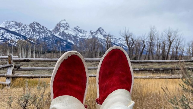 Feet in red moccasins held up to a grassy field with a mountain range in the distance 