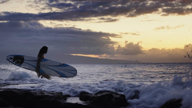 Person holding a surfboard while standing in ocean waves, text reads "Koa talking to me"