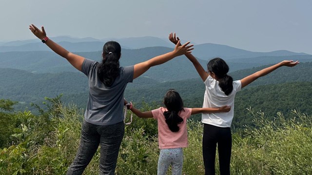 A woman and two girls hold their hands open above their hands at an mountain overlook.