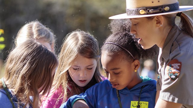 A woman in a flat-brimmed tan hat and gray uniform shirt holds an item for four young children to lo