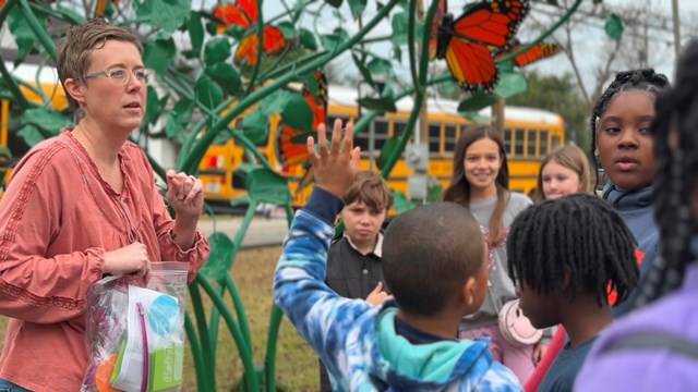 A teacher and six students next to a butterfly sculpture with a yellow school bus in the background.