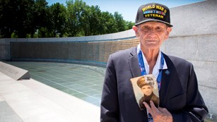 World War II veteran holding a black and white photo of himself while standing in a memorial