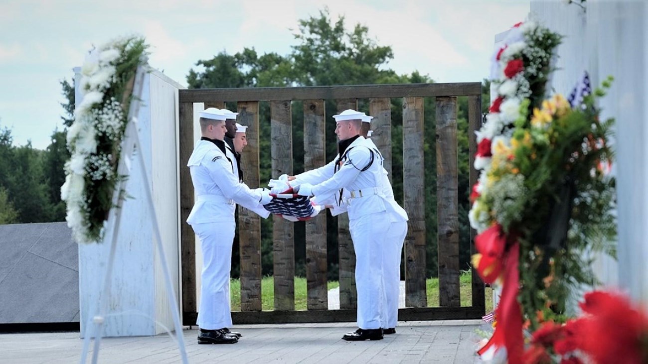 Military service members folding a flag near a wreath and memorial wall