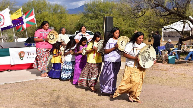 7 women and girls in skirts hold baskets in front of a stage at an event.