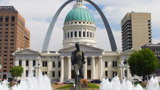 A statue in a fountain in front of a white courthouse backed by a large silver arch.