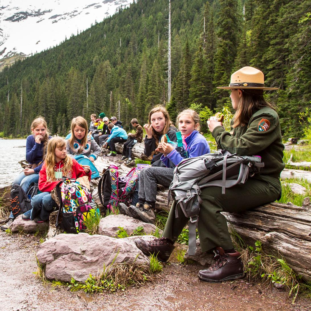A ranger and students sit on a log near a lake with mountains the background