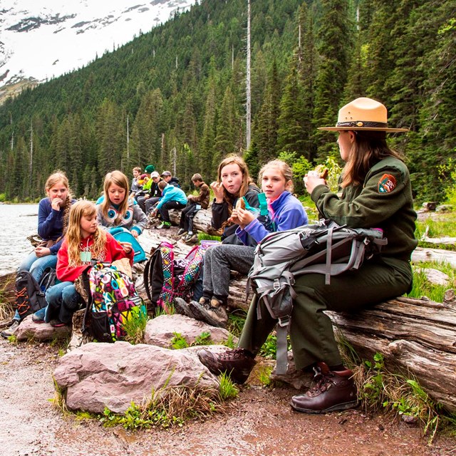 A ranger and students sit on a log near a lake with mountains the background