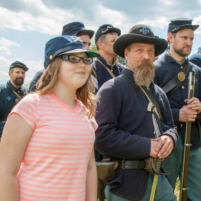 Child wearing a military hat while standing next to a line of Civil War reenactors