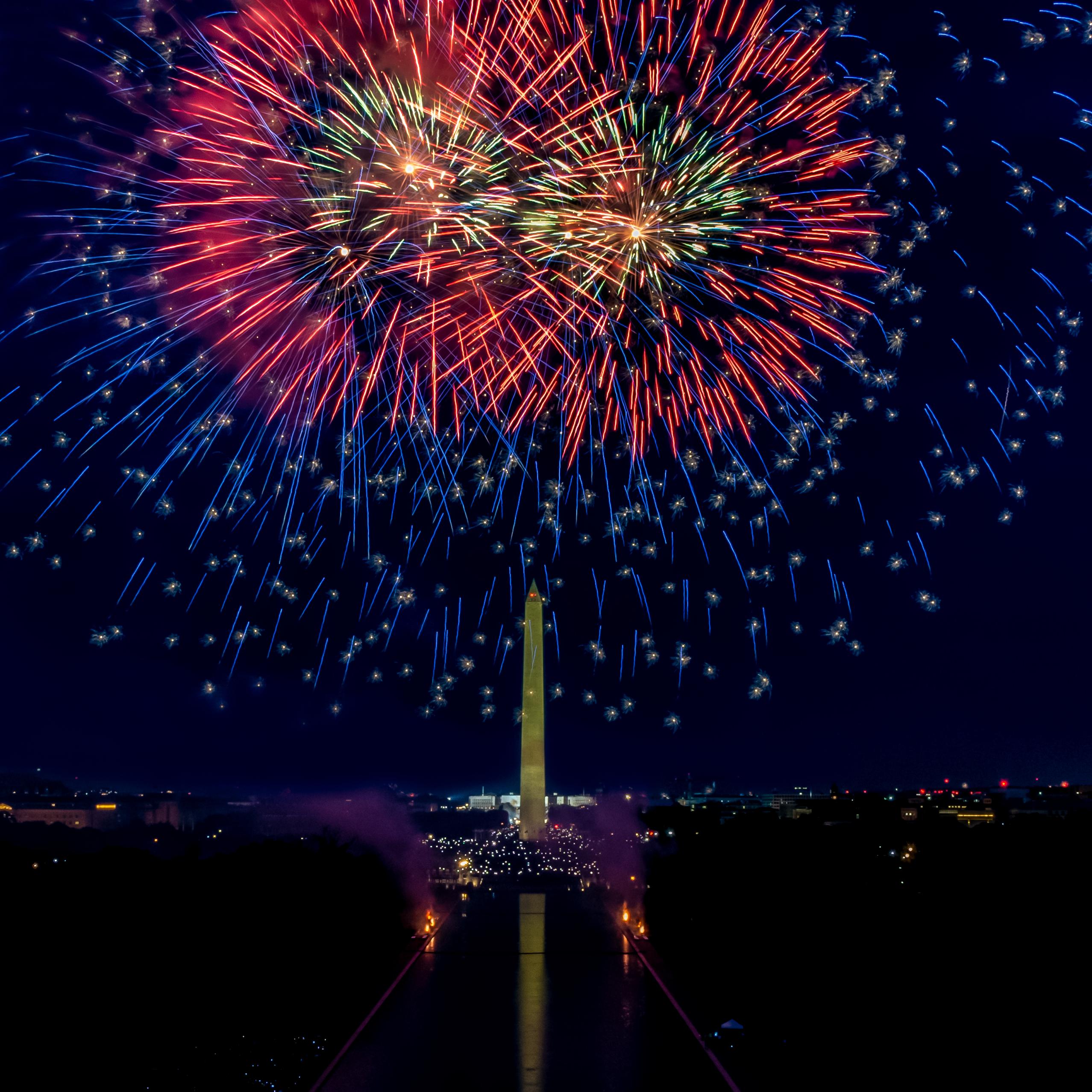 Red and blue fireworks exploding in a dark sky above the Washington Monument and Reflecting Pool