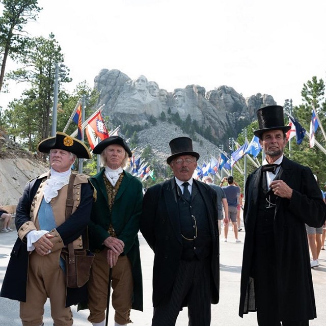 four men dressed up in founding fathers clothing with mount rushmore in the background
