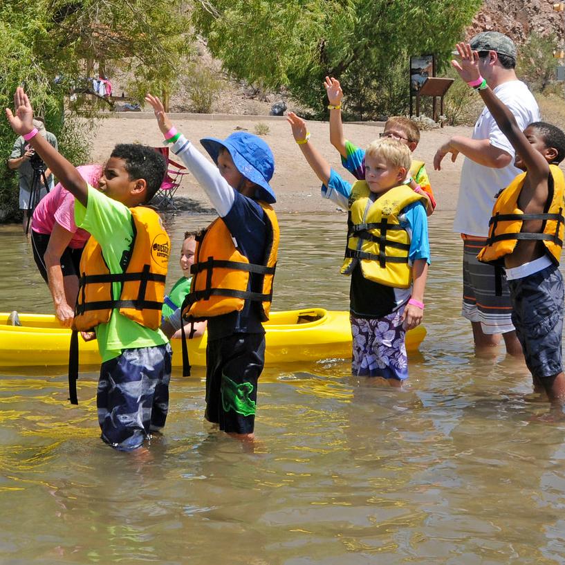 A group of kids standing in the water next to a kayak interacting with an instructor