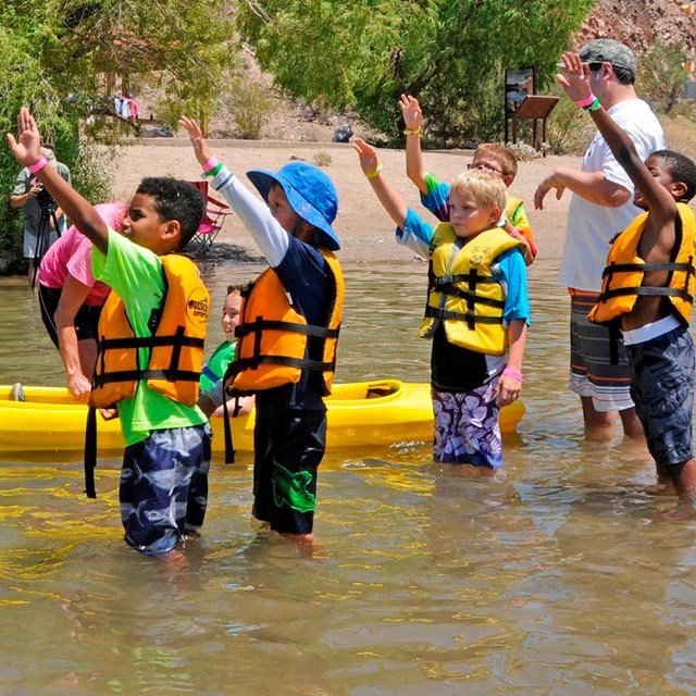 A group of kids standing in the water next to a kayak interacting with an instructor