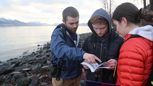 Three young people look through a field guide on a rocky coastline
