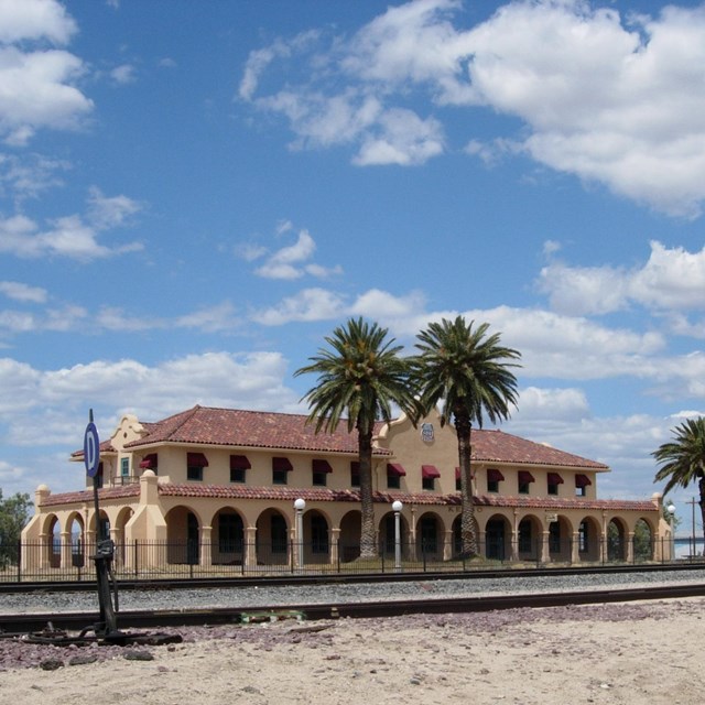 A long one story building with a covered walkway, set in the desert next to some palm trees.