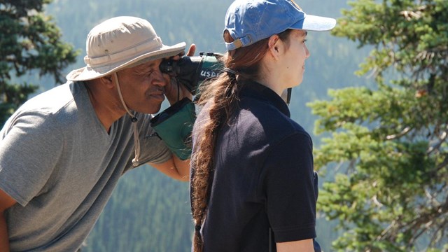 A man looks through a scope. A woman stands next to him.